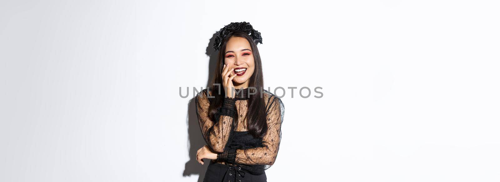 Image of happy good-looking asian woman in gothic dress and black wreath, laughing at halloween party, standing over white background.