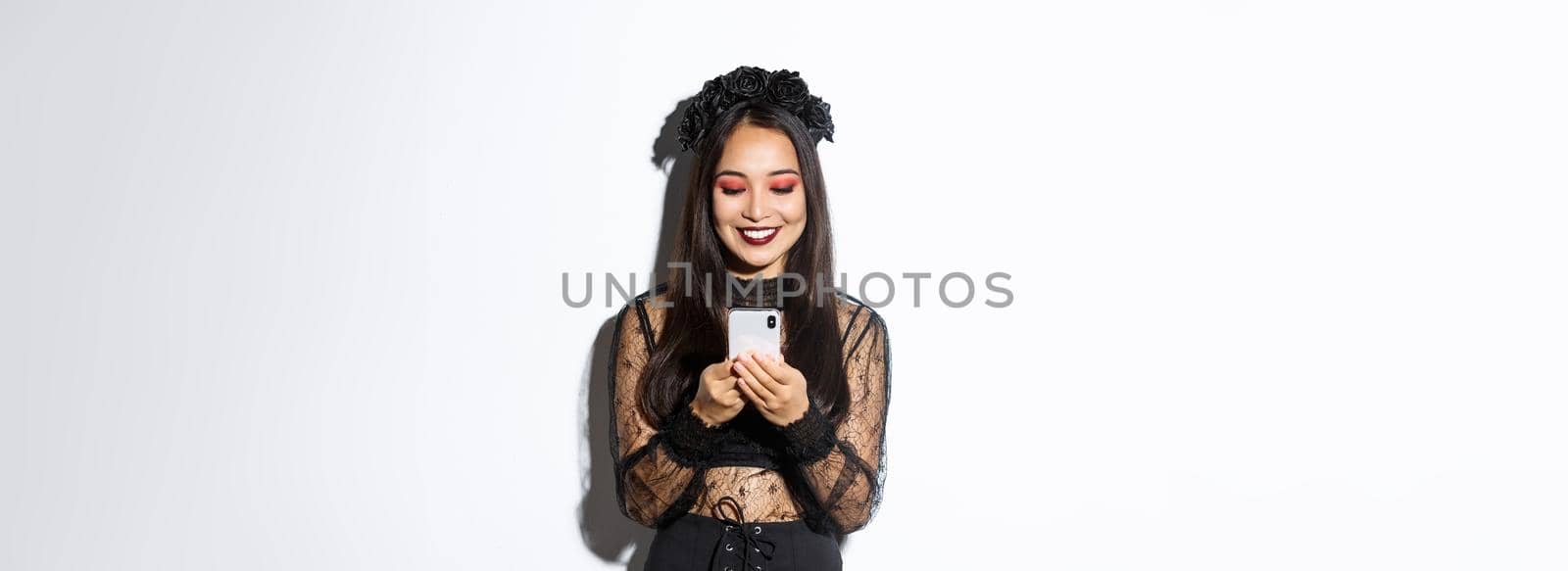 Image of beautiful asian woman in gothic lace dress impersonating witch on halloween party, standing with smartphone and smiling over white background.