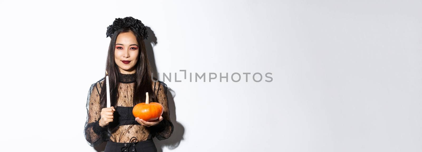 Image of devious smiling asian witch in gothic dress, holding candle with pumpkin and looking at camera cunning, standing over white background.