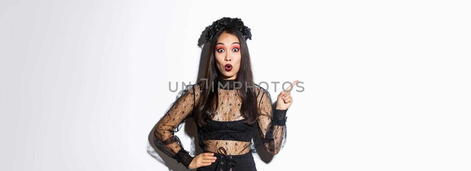 Surprised attractive asian woman in witch outfit pointing finger at upper left corner, showing halloween advertisement, promo of party. Beautiful female in black gothic dress looking amazed.