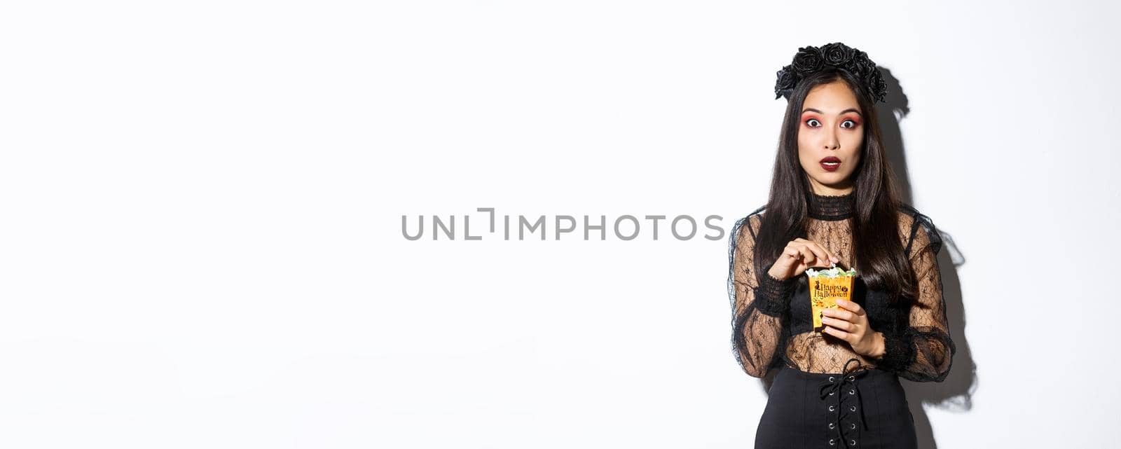 Image of surprised and fascinated asian woman in witch costume, holding sweets gathered during trick or treating, standing over white background.