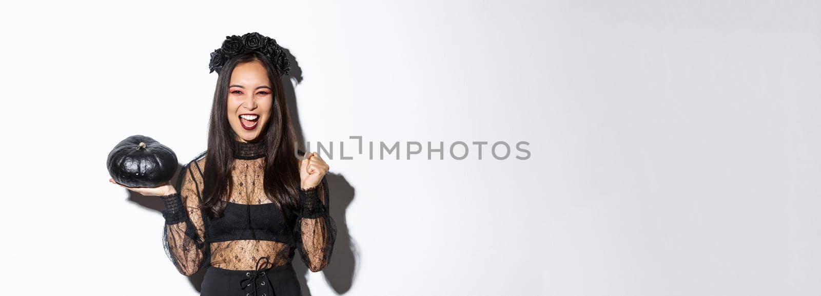 Attractive joyful asian girl enjoying halloween, holding black pumpkin and cheering, wearing witch costume, standing over white background.