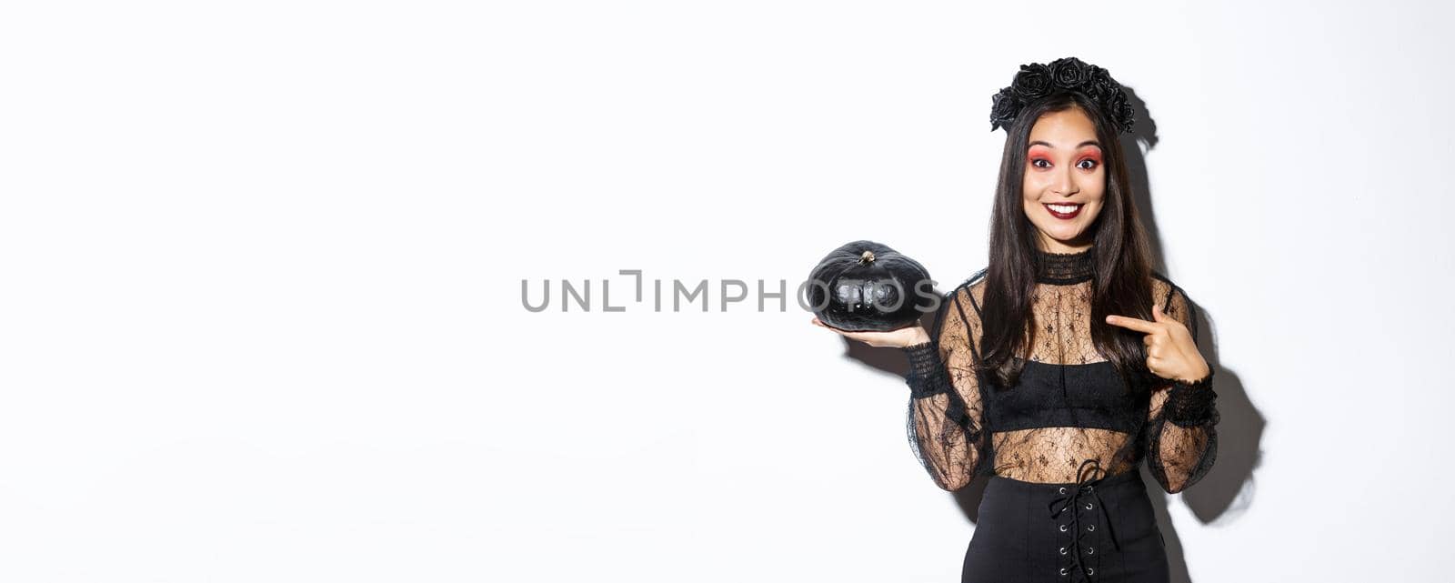 Surprised happy asian woman in witch costume pointing finger at black pumpkin, smiling amused, getting ready for halloween party, standing over white background.