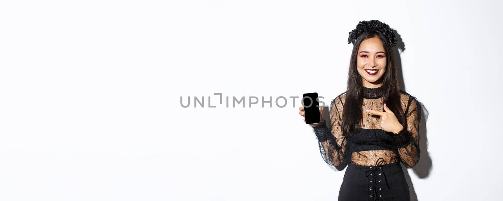 Happy beautiful asian girl in witch costume pointing finger at smartphone screen with pleased smile, showing halloween announcement, white background.