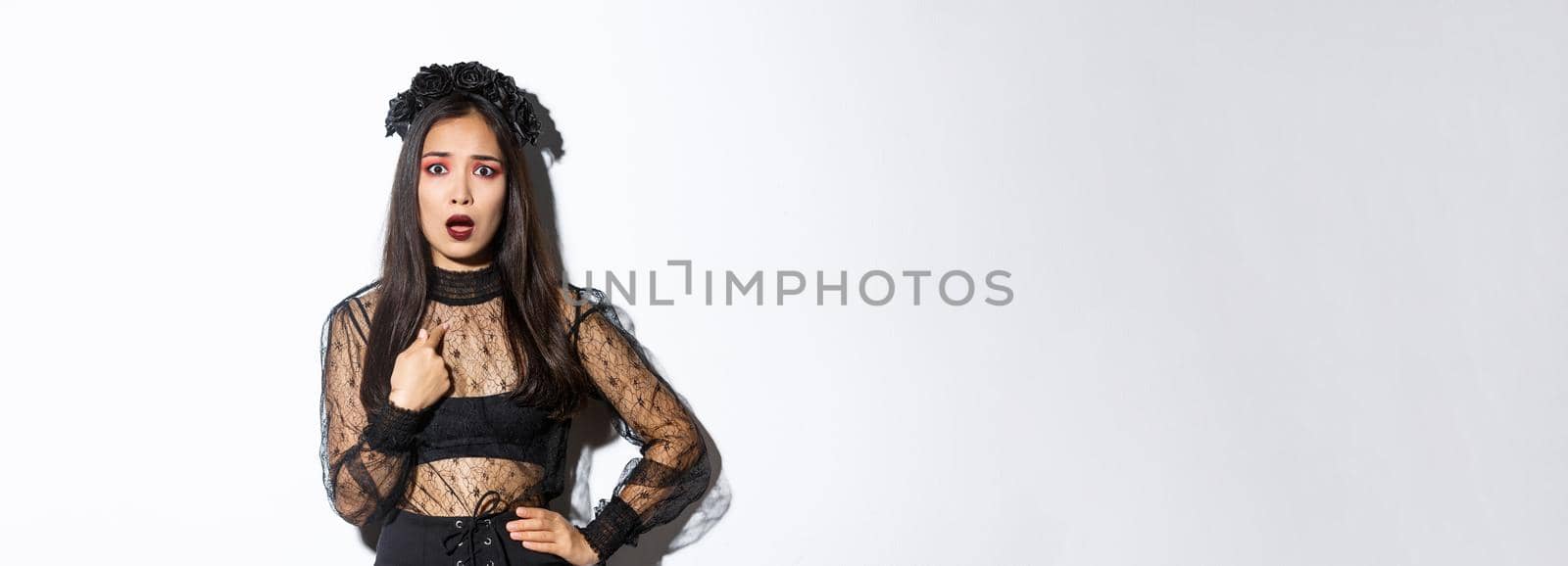 Insulted and confused asian woman in halloween costume pointing at herself, wearing black dress of evil witch on party, standing over white background.