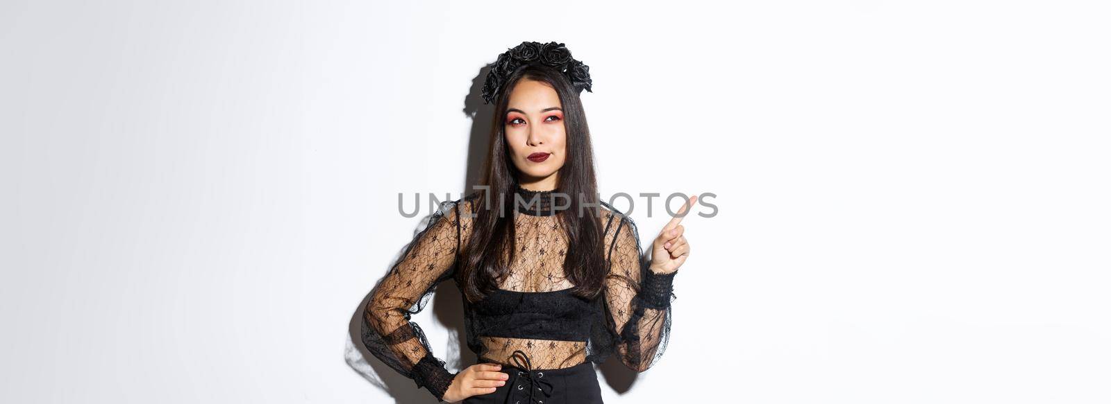 Curious attractive asian woman in witch costume, pointing finger upper left corner, looking interested in halloween promo, standing over white background and thinking.