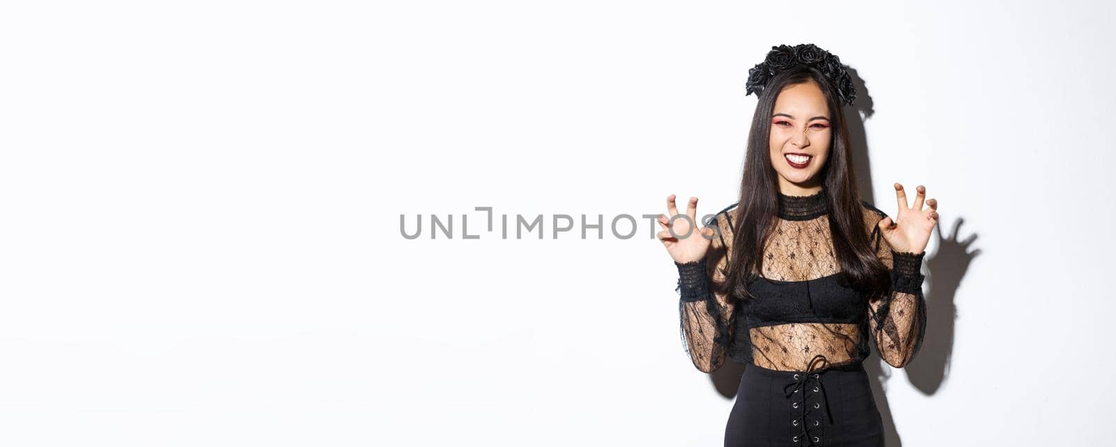 Sassy beautiful asian woman in black lace dress and wreath trying to scare you, raising hands up, acting like evil witch on halloween trick or treat event, standing over white background.