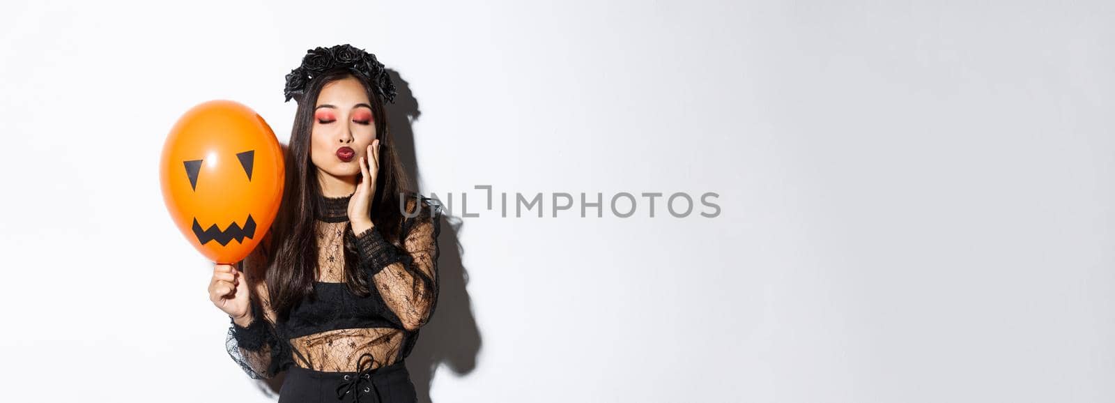 Image of beautiful stylish asian woman celebrating halloween, wearing witch costume, holding orange balloon with face.