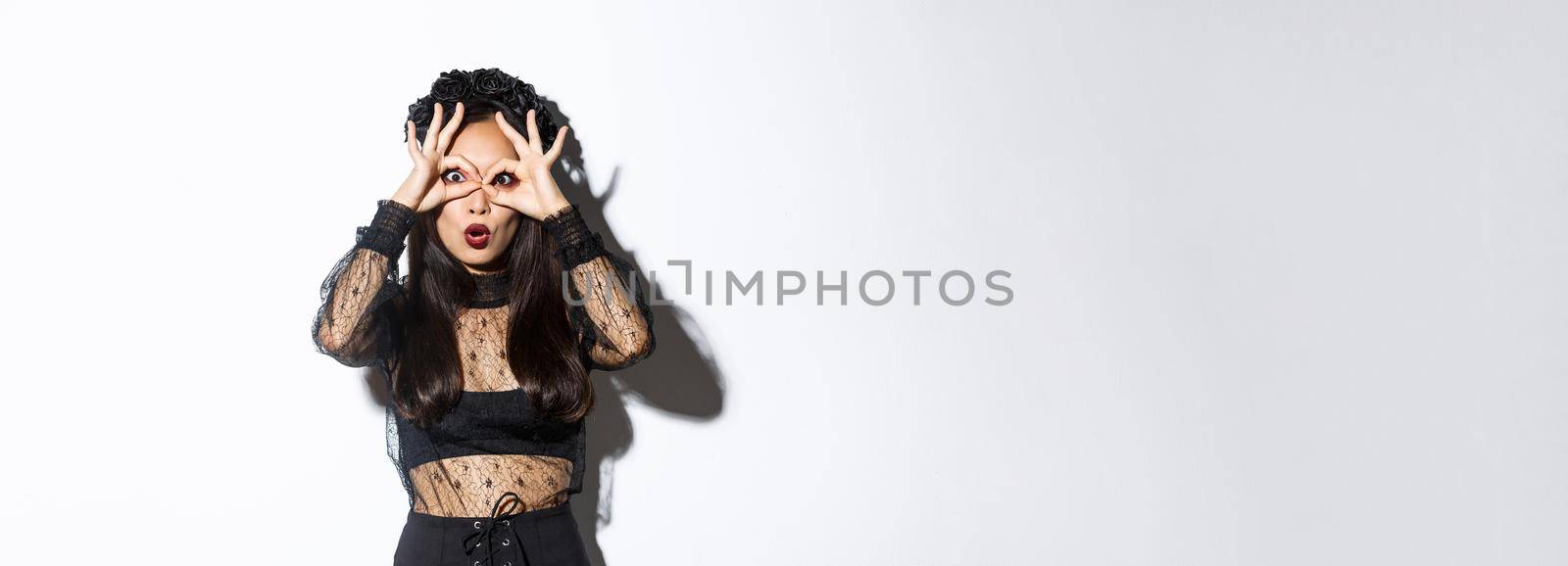 Image of surprised cute asian woman wearing witch costume, making finger glasses over eyes and looking through them impressed, standing over white background.