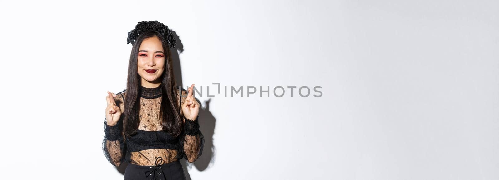 Image of happy asian girl who loves celebrating halloween dressed-up as witch, smiling hopeful and cross fingers for good luck, making wish, standing over white background.