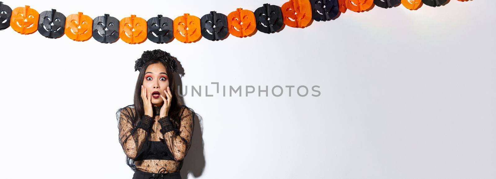 Image of woman in witch costume looking scared, express horror or fear while standing over white background with pumpkin banners decoration, celebrating halloween.