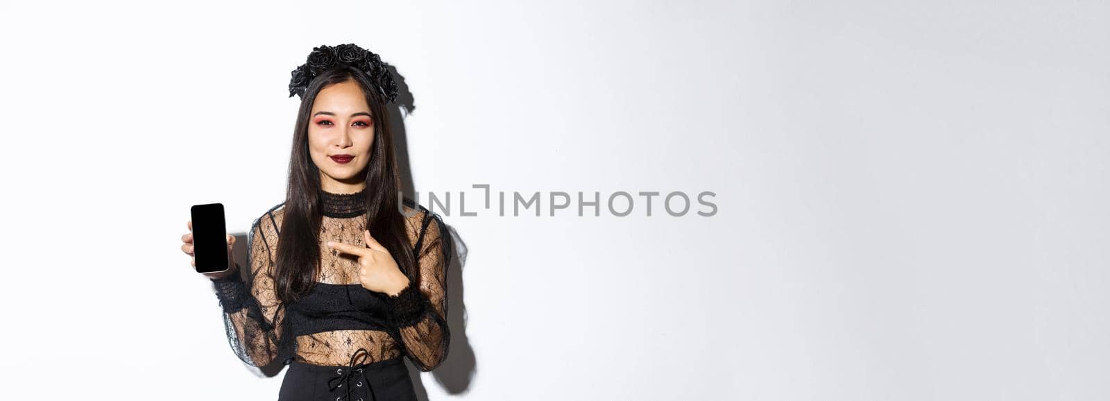 Young elegant woman in gothic dress and black wreath pointng finger at smartphone screen with pleased smile on her face, standing over white background by Benzoix