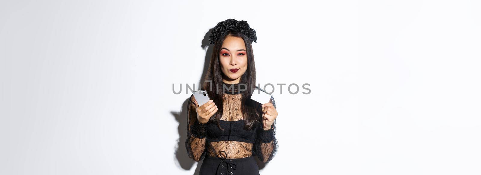 Sassy young woman looking thoughtful, holding credit card and mobile phone, shopping in internet, standing over white background.