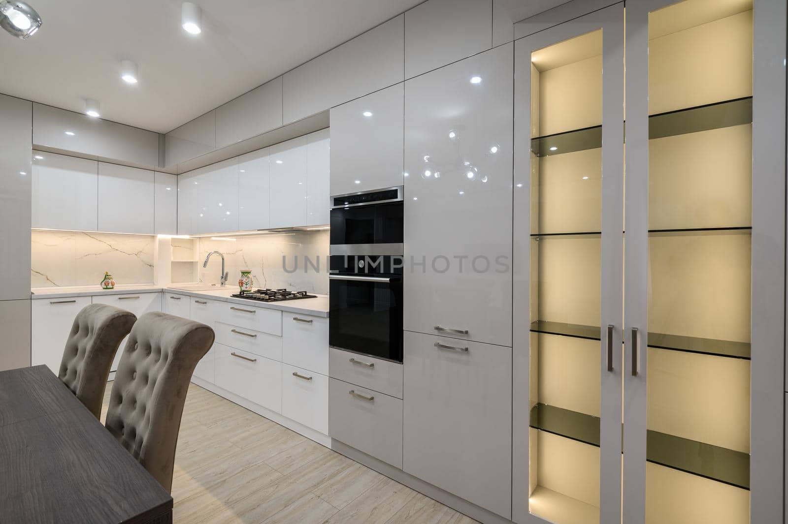 Dining room table and chairs, white spacious furnished kitchen interior with oven and large LED enlightened sideboard at right side