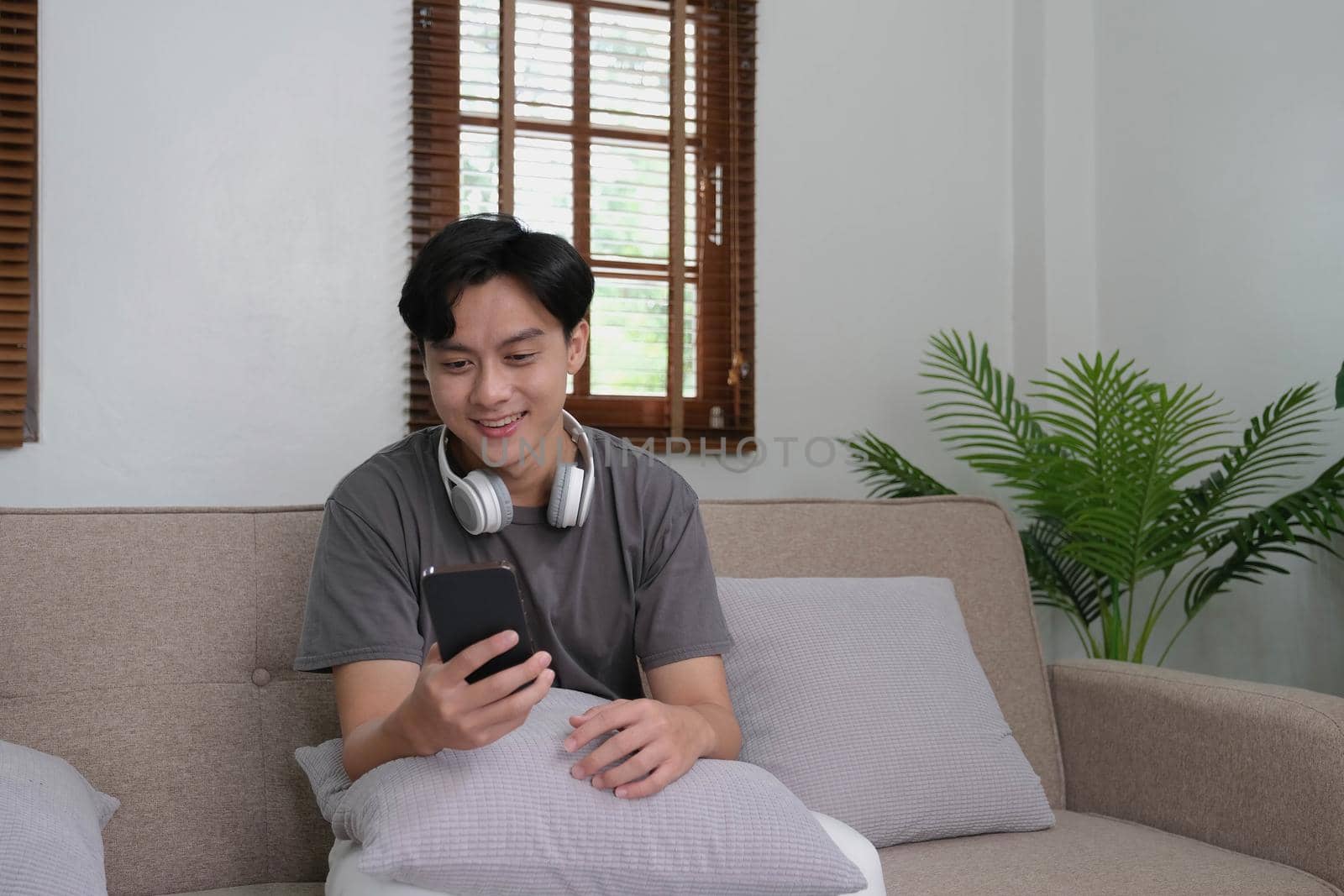 Handsome Asian man using smartphone while headphones sitting on sofa at home.