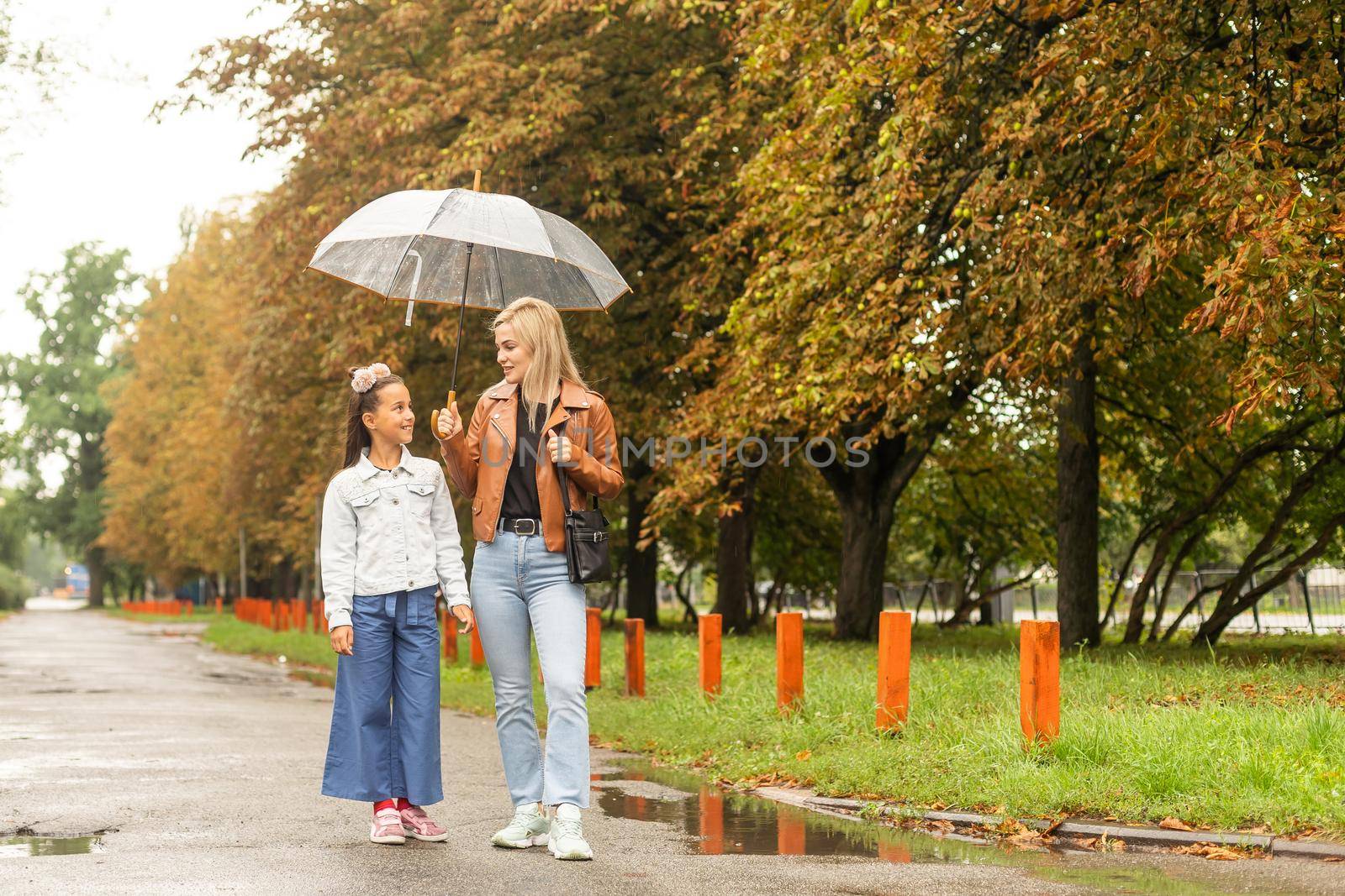 Mom holds a transparent umbrella from which fall autumn leaves. Daughter and mother laugh. Happy mom and little daughter having fun in autumn park