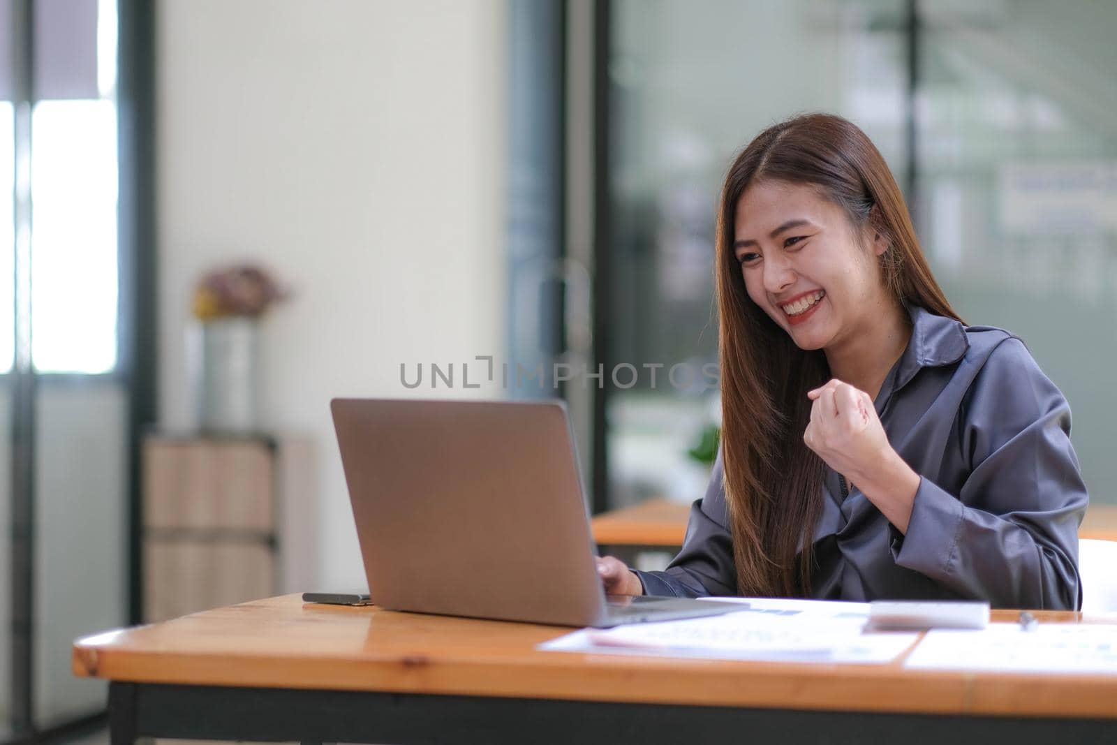 Happy excited successful Asian businesswoman triumphing with a laptop computer smartphone in the workplace office.