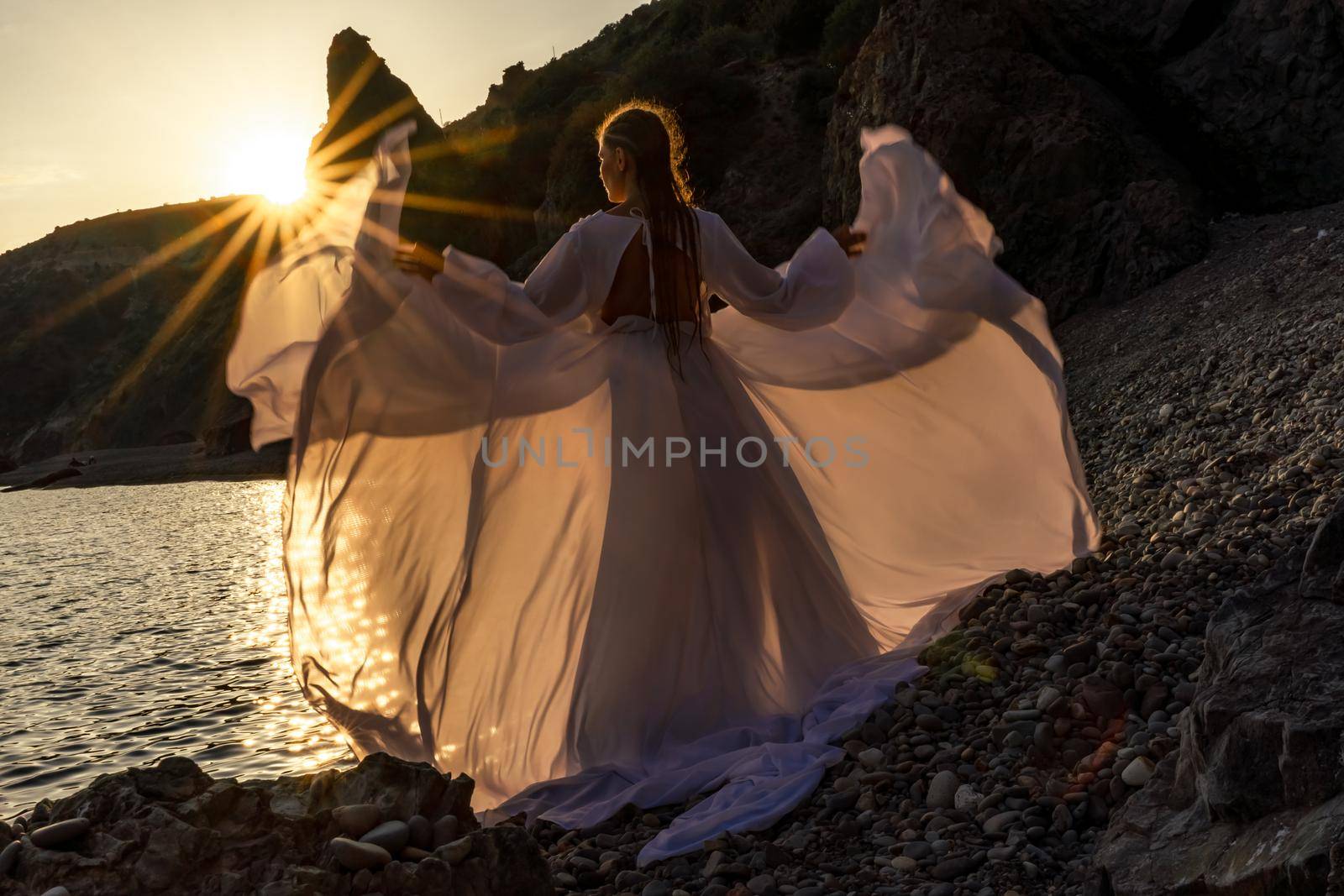 A mysterious female silhouette with long braids stands on the sea beach with mountain views, Sunset rays shine on a woman. Throws up a long white dress, a divine sunset