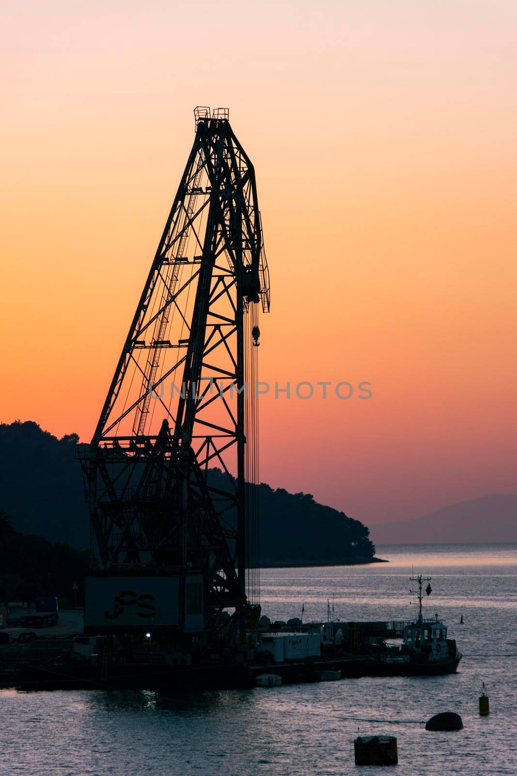 Portrait photo of black, dramatic silhouette of industrial crane against a gradient of pink, orange and purple clear sky, with sea water in foreground on a beautiful island in croatia