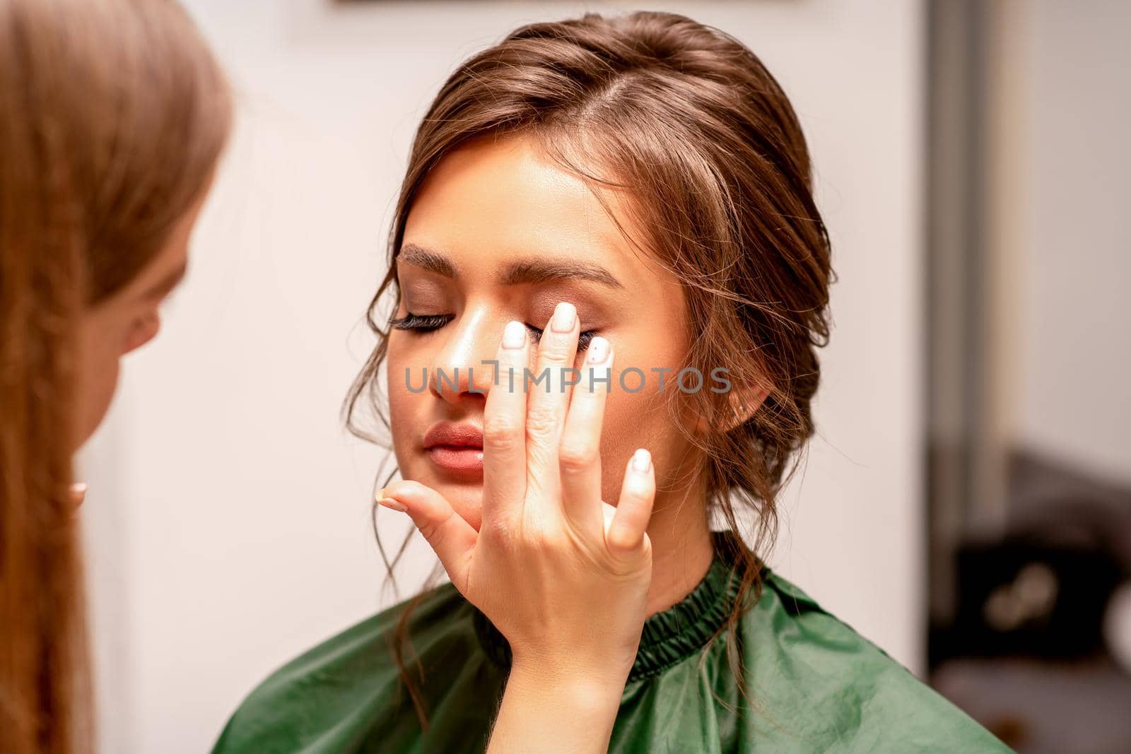 The hand of a makeup artist applies eye shadow on the eyelid of a young Caucasian woman with fingers in a beauty salon