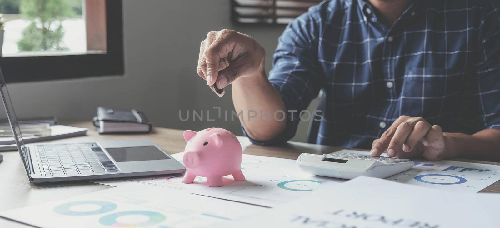 Portrait of an Asian businessman using a calculator to calculate his savings from SME operations, with a pink piggy bank as keep money concept..