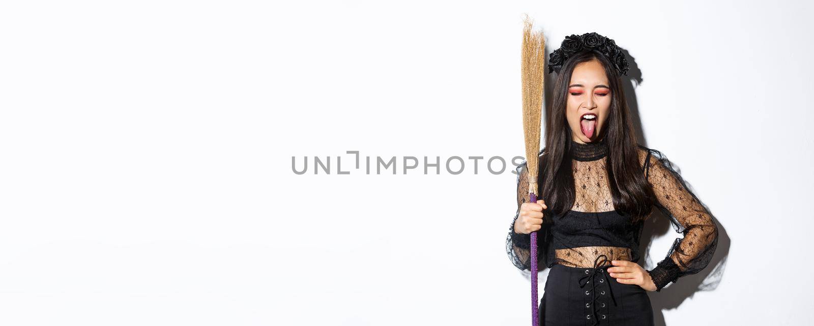 Carefree sassy asian woman enjoying halloween party, wearing witch costume and holding broom, showing tongue, standing over white background.