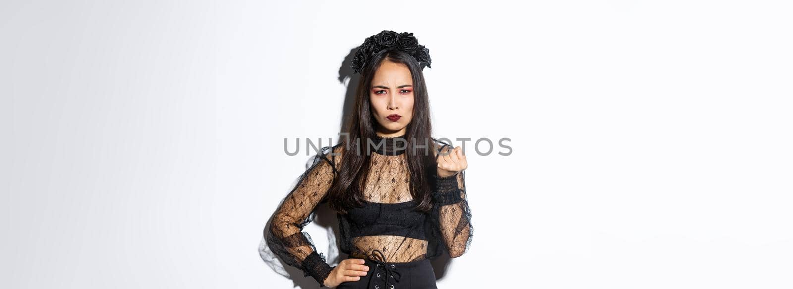 Image of woman wearing evil witch costume for halloween and threaten someone with fist. Girl in black gothic dress and wreat scolding person, looking angry, standing white background by Benzoix
