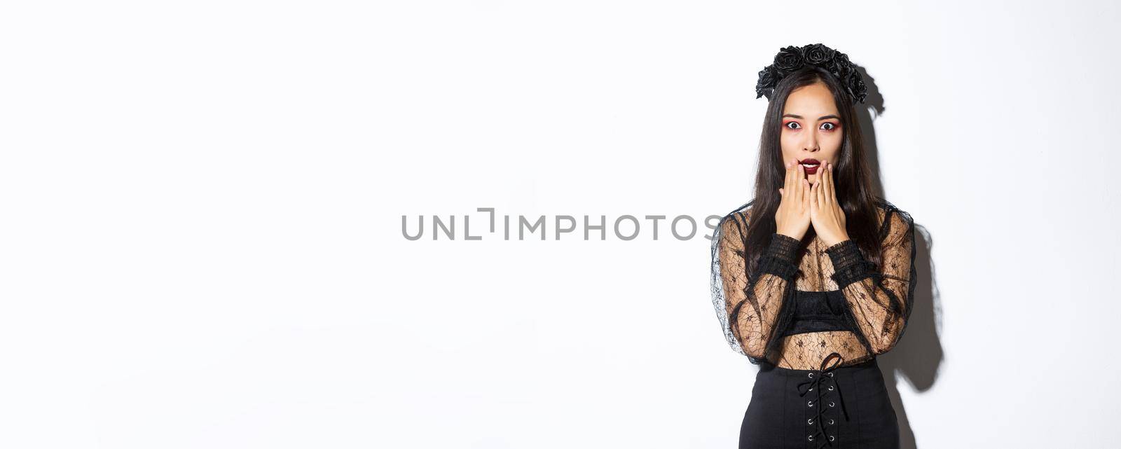 Shocked and startled asian woman, witch in black lace dress looking concerned, gasping and looking at camera amazed, standing over white background.