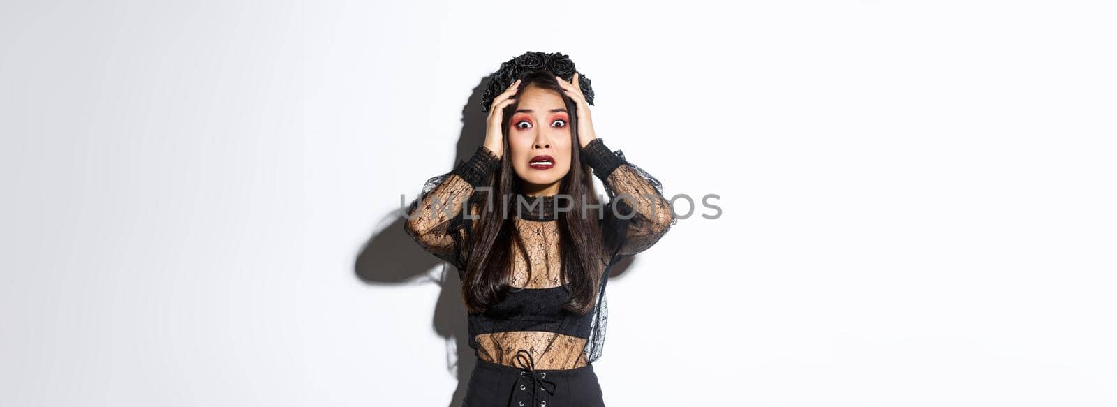 Image of scared and worried asian woman in halloween costume looking horrified or anxious, holding hands on head and stare at camera, panicking over white background.