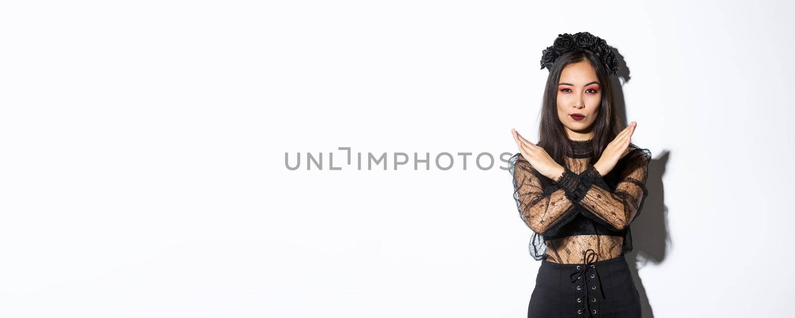 Confident beautiful woman in black gothic dress showing cross gesture, disapprove and stop something bad, disagree with someone about halloween, standing over white background.