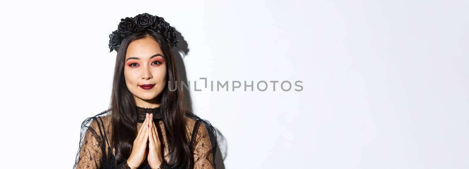 Close-up of young asian woman in black gothic dress and wreath holding hands in pray, girl wearing witch costume and celebrating halloween.