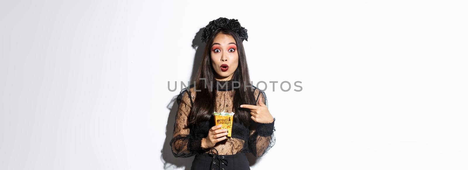 Amazed asian girl looking at camera while pointing finger at sweets, gather treats on halloween, wearing witch dress, standing over white background.