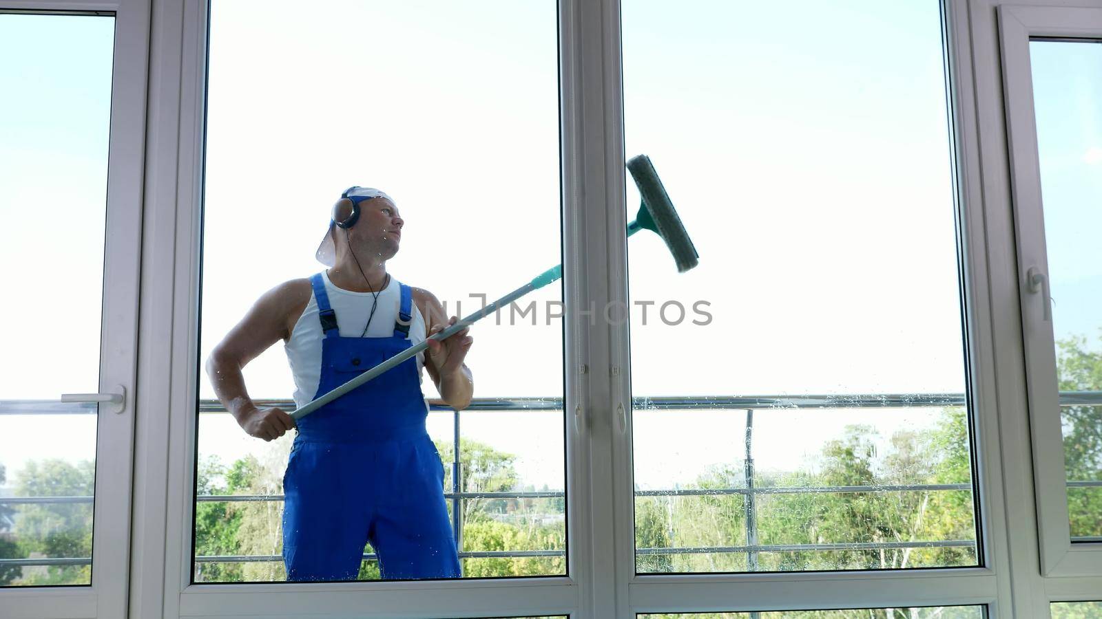 view through window,handsome male worker of cleaning service, in blue overalls, with headphones, , Cleaning, washing Windows by special mop, enjoying his work and music. High quality photo