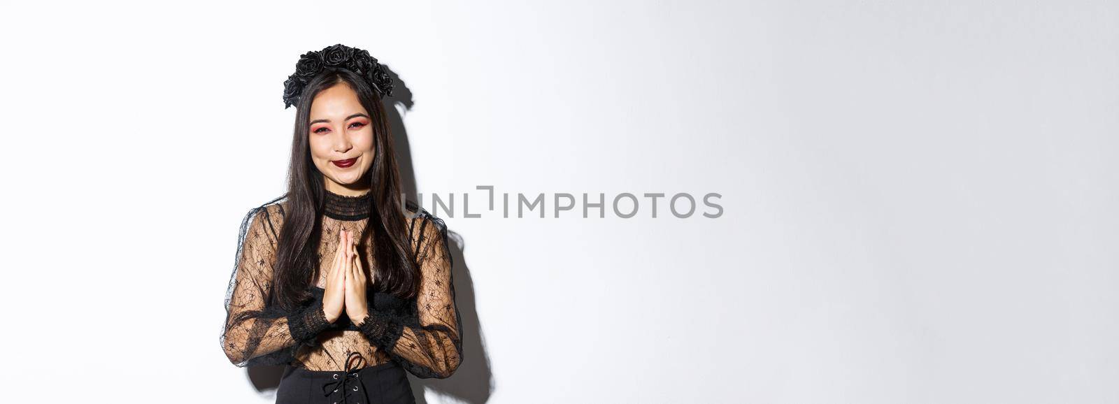 Image of happy and grateful young asian woman in gothic lace dress clasp hands together to say thank you, smiling thankful and standing over white background by Benzoix