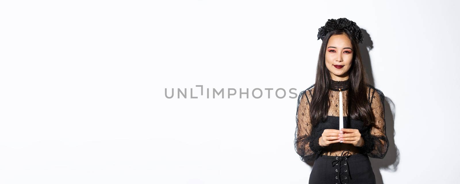 Image of beautiful woman celebrating halloween in witch costume, holding candle and squinting at camera suspicious, standing over white background.