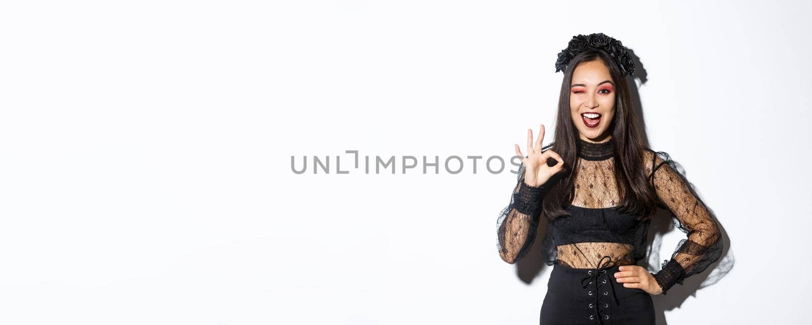 Attractive young asian woman in halloween gothic dress, showing okay gesture, give permission, approve or like your party costume, standing pleased over white background by Benzoix