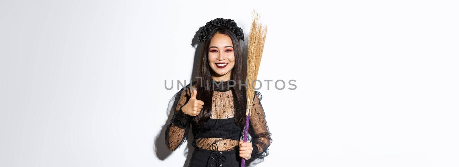 Image of smiling asian woman in witch costume with broom, showing thumbs-up in approval, standing over white background.