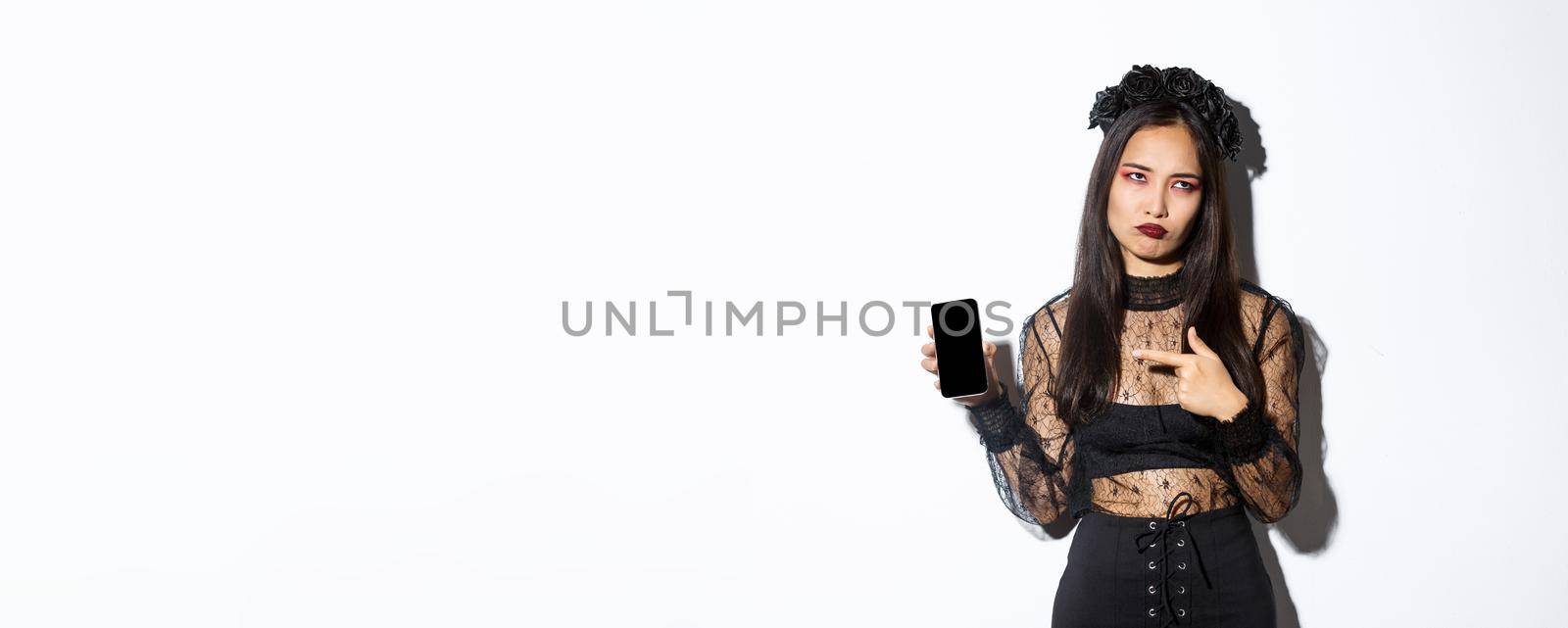 Image of annoyed and reluctant asian girl in witch costume, roll eyes and pouting while pointing finger at mobile phone screen, standing over white background.