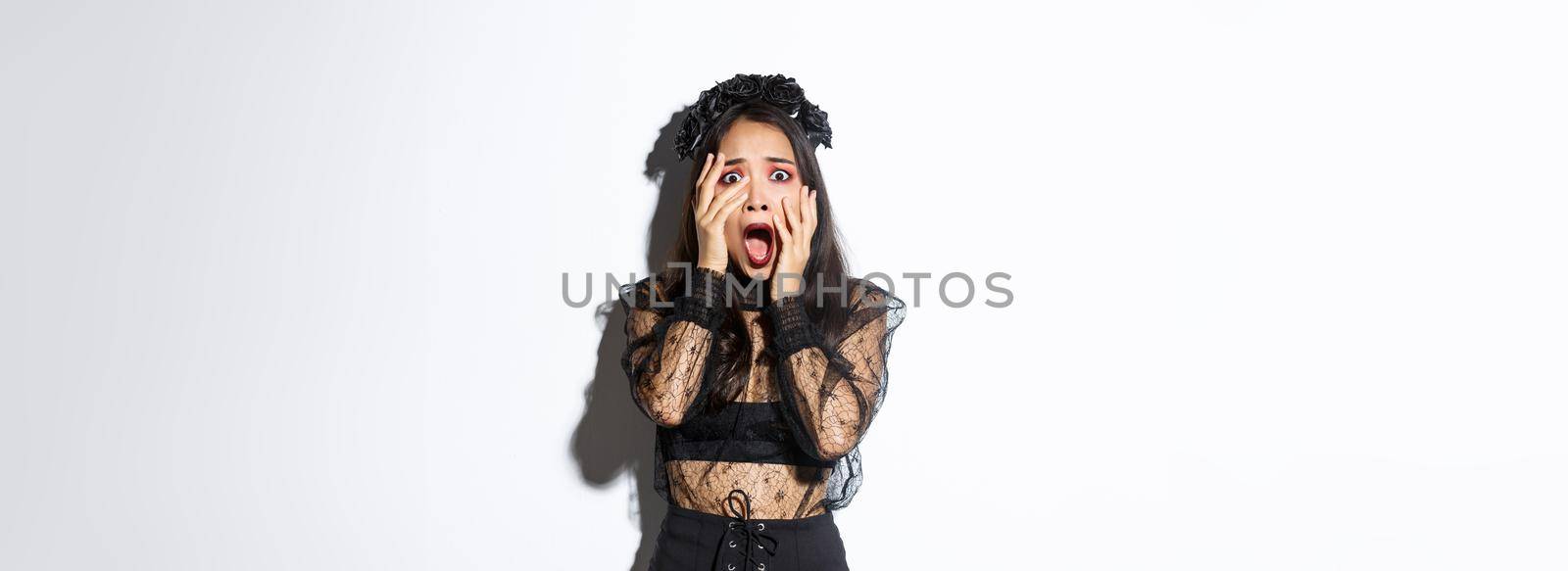 Image of scared young woman screaming and looking frightened at something creepy on halloween, wearing gothic dress and wreath, holding hands on face and trembling from fear.