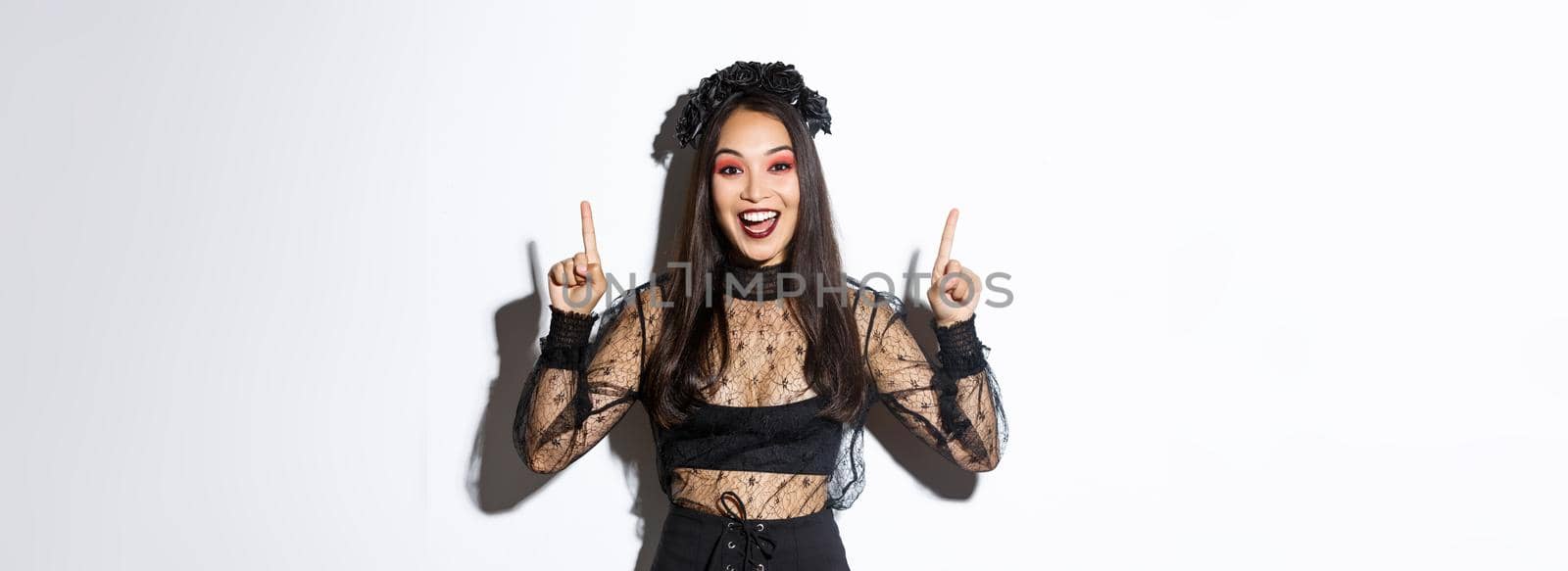 Excited smiling woman celebrating halloween in black gothic dress and wreat. Asian girl wearing witch costume and pointing fingers up, standing white background.