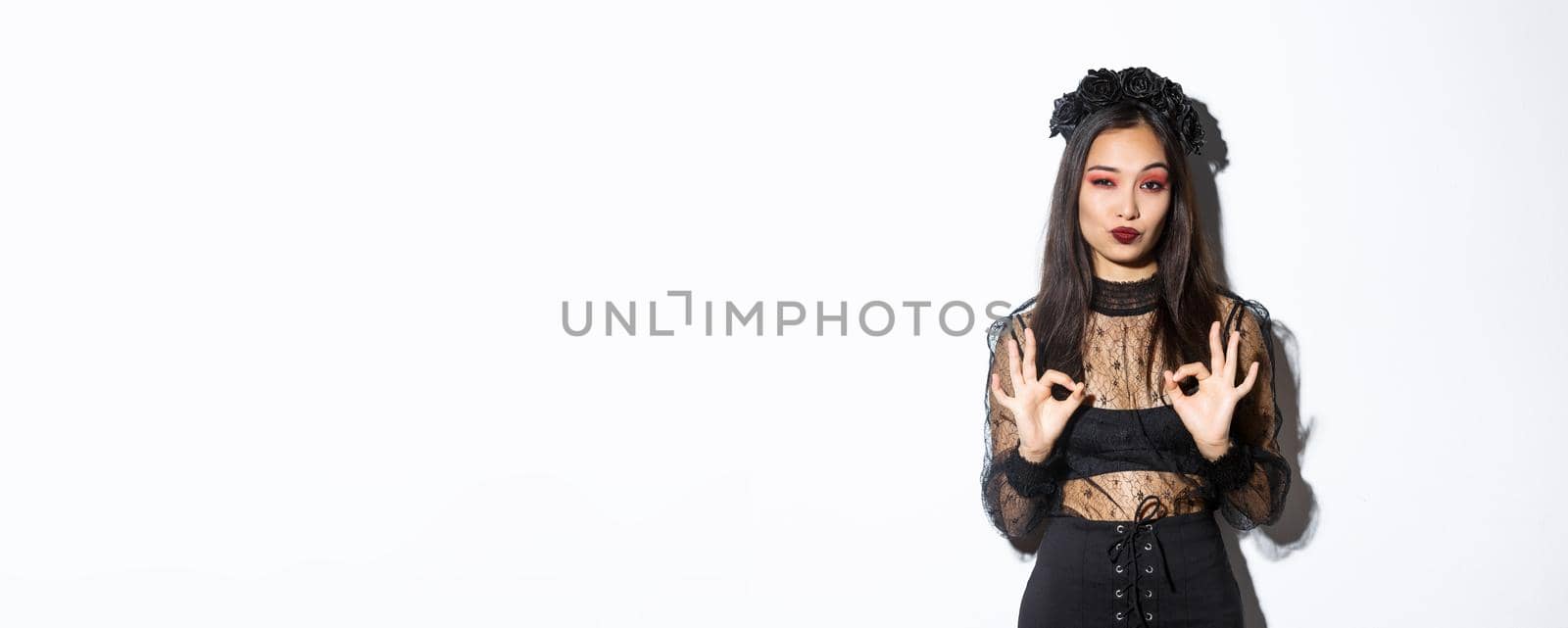 Image of stylish asian woman in gothic dress, looking satisfied and showing okay gestures in approval, praise something good, standing impressed over white background.