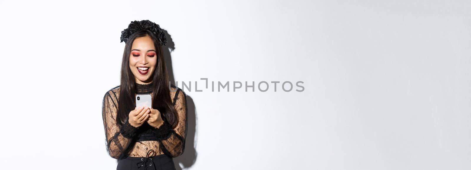 Excited smiling woman looking at mobile phone pleased, wearing gothic lace dress for halloween party, standing over white background.