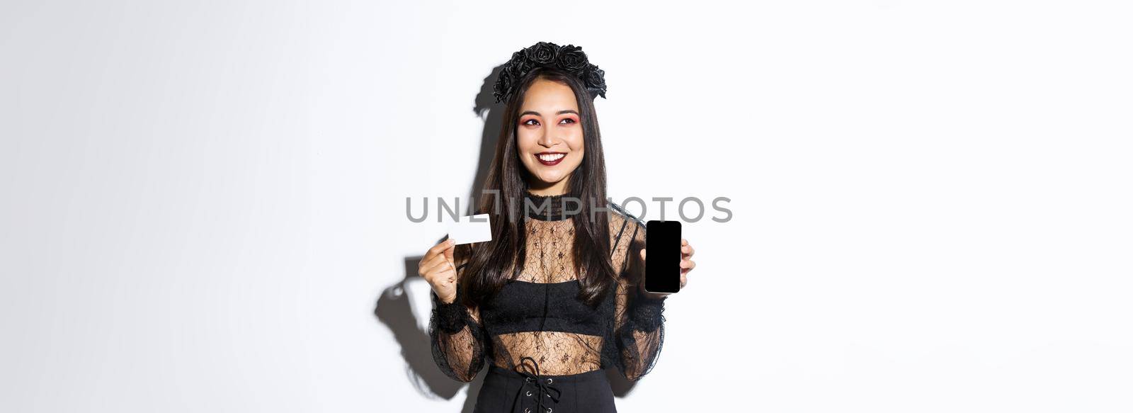 Dreamy smiling girl looking away while thinking, showing credit card and mobile phone, wearing halloween gothic dress by Benzoix