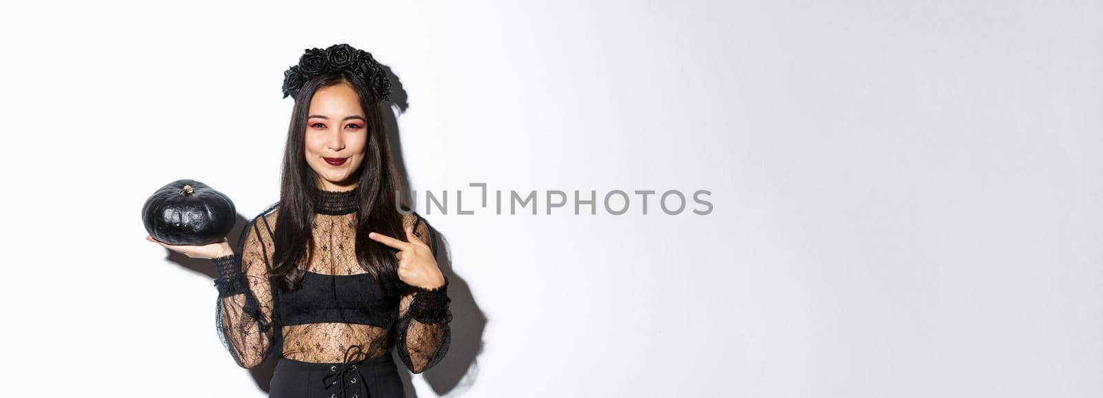 Image of beautiful asian woman in gothic lace dress and wreath pointing finger at black pumpkin, celebrating halloween, wearing witch costume, standing over white background by Benzoix