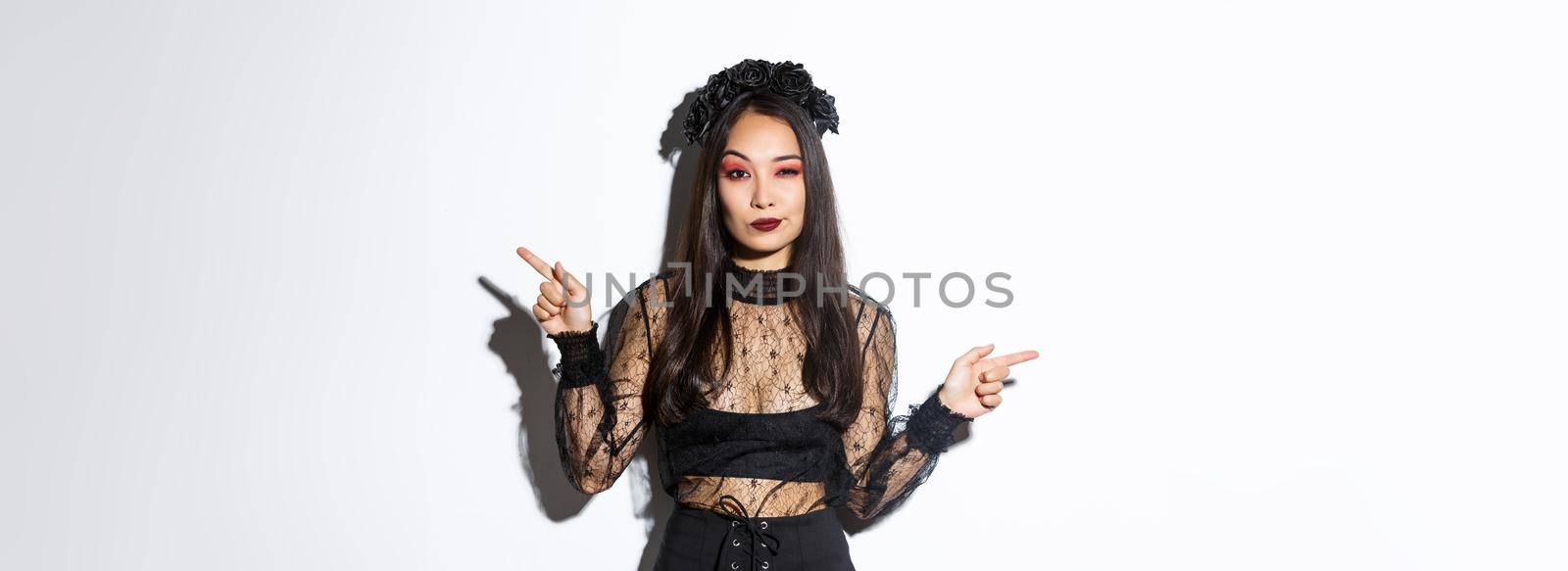Sassy young evil witch with gothic makeup and wreath, looking arrogant while pointing fingers sideways, showing two halloween themed banners, standing over white background.