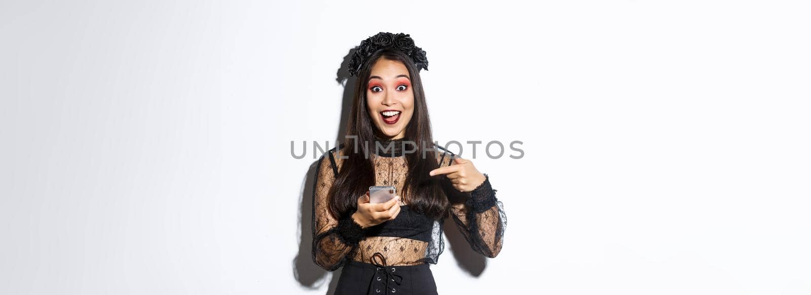 Portrait of excited asian girl pointing finger at smartphone, showing something about halloween in internet, standing over white background in gothic lace dress.