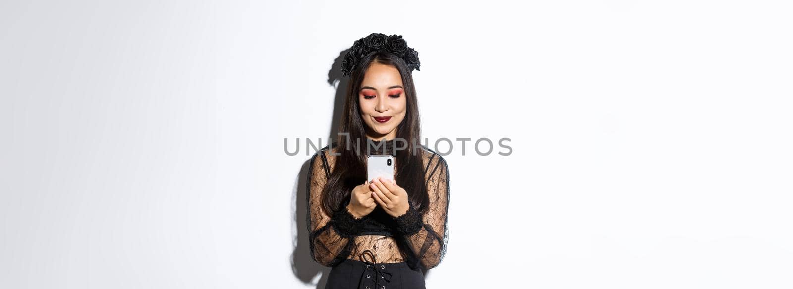 Image of stylish asian woman in halloween costume checking messages on mobile phone. Girl in gothic lace dress looking at smartphone, standing over white background.
