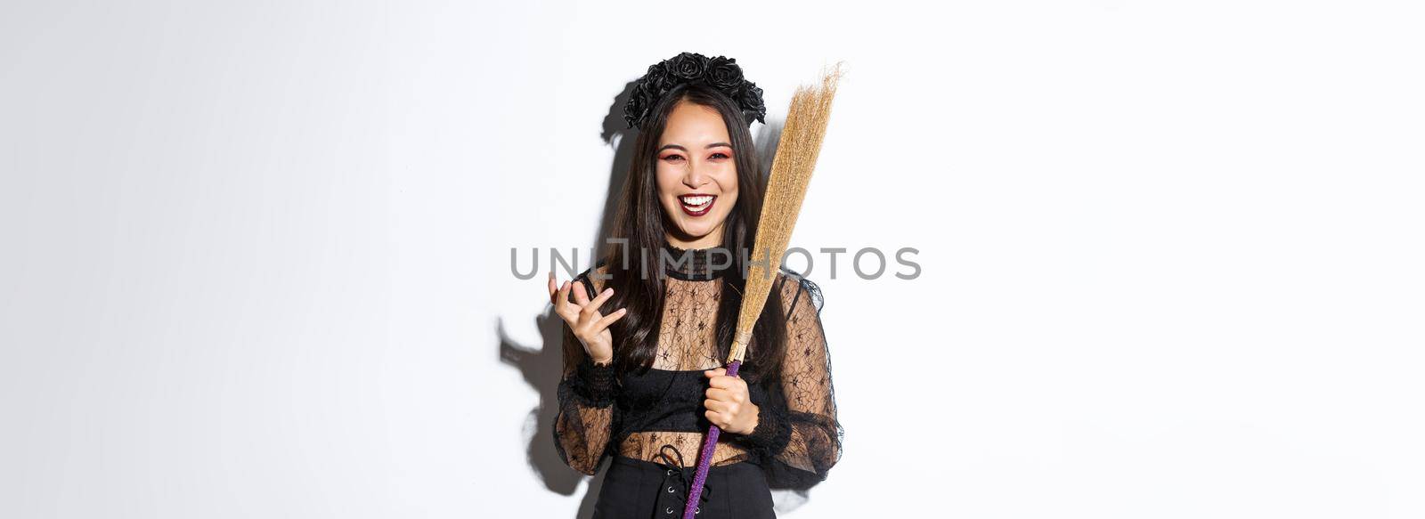 Image of witch in gothic lace dress and black wreath making evil laugh and holding broom, celebrating halloween, standing over white background.