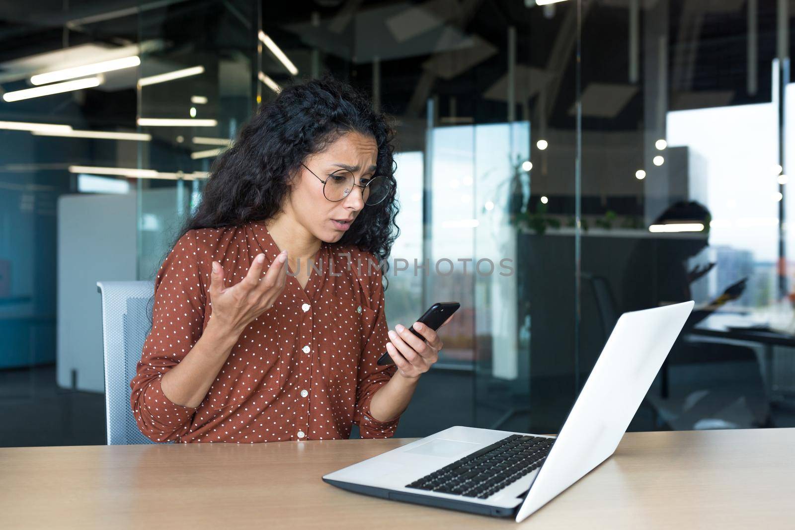 Disappointed and sad woman looking at phone screen, reading bad news online, Hispanic business woman working inside modern office building.