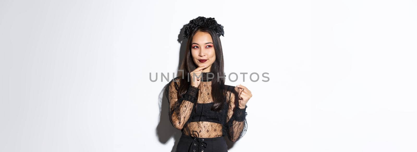 Image of smiling beautiful asian woman in gothic lace dress and wreath, thinking while holding credit card, standing over white background.